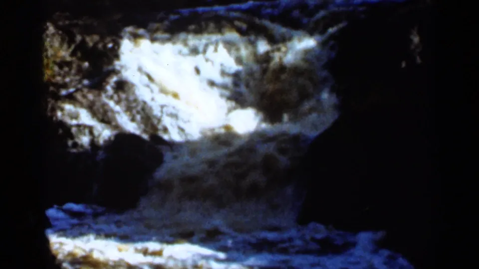 1965 Duluth home-movie frame showing turbulent white water moving over rocks in a dark, shaded setting.