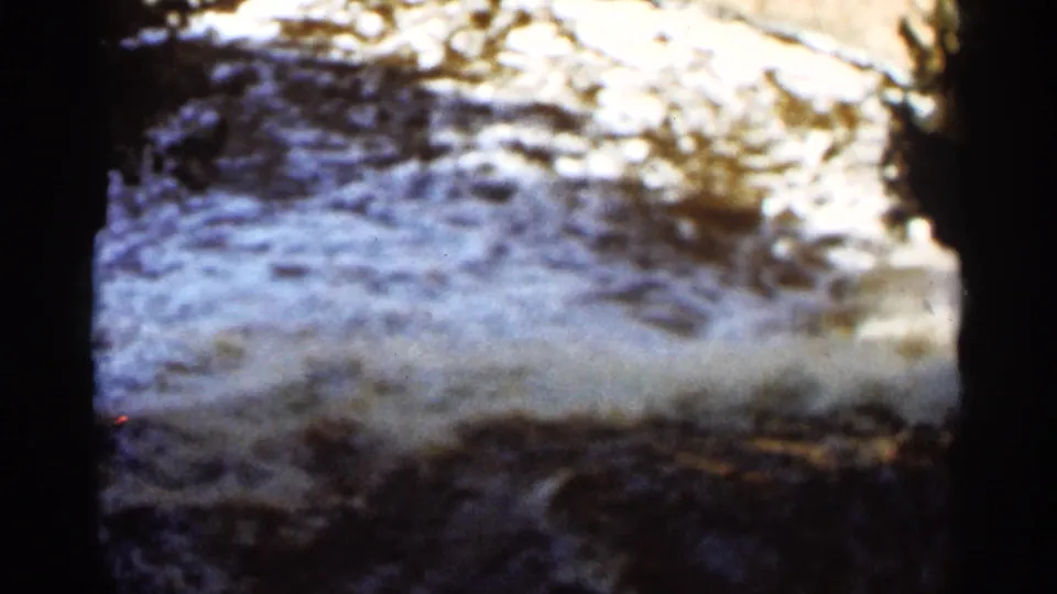 1965 home-movie frame of bright rippling creek water over rocks, softened by 8mm grain and color shift.