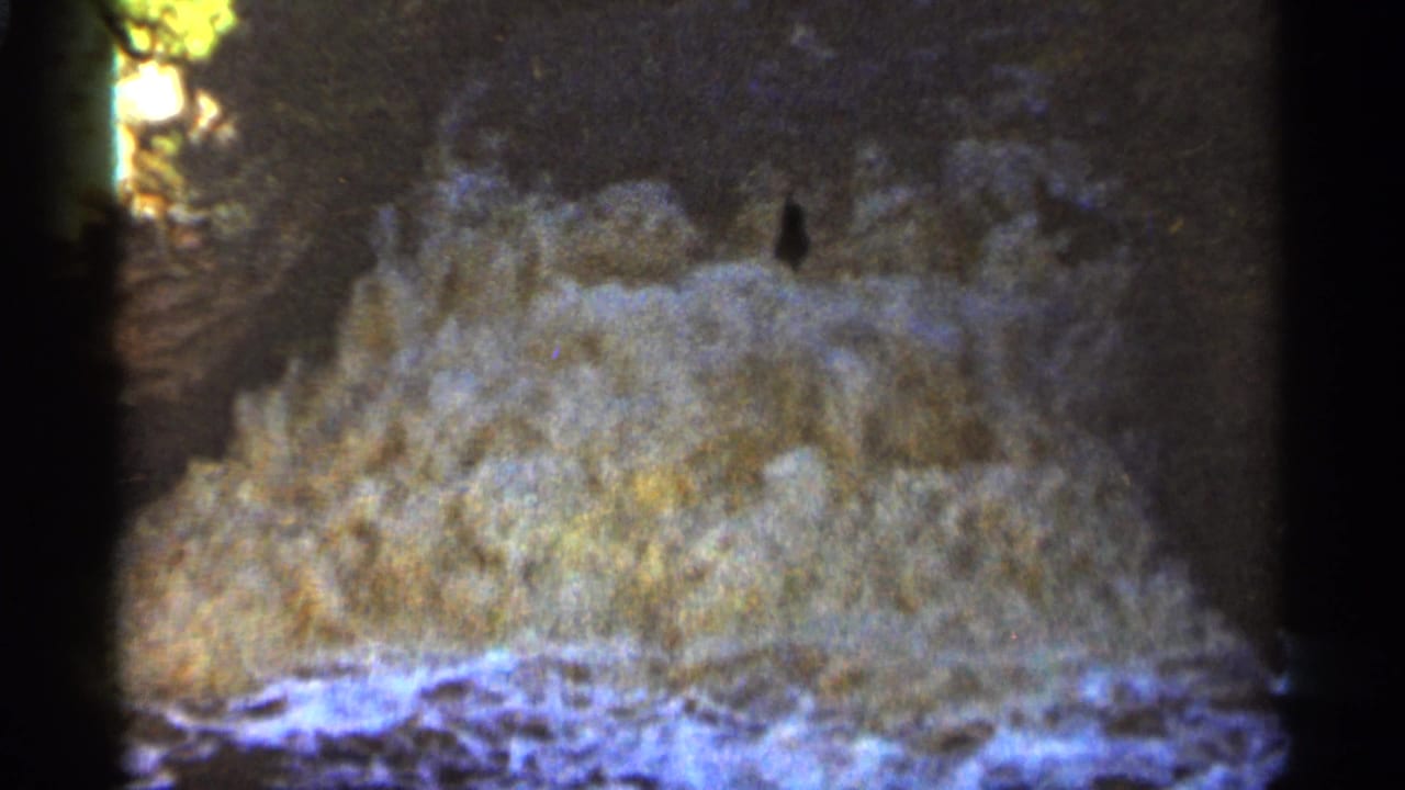 Foamy water crashes over dark rocks in a grainy 8mm frame.
