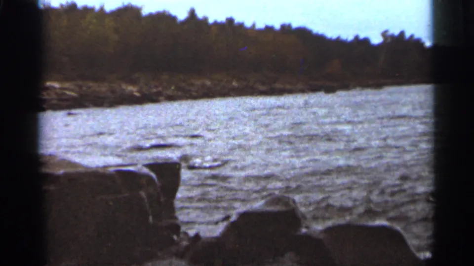 A wider Duluth shoreline view shows rippled water, foreground rocks, and a wooded opposite bank in 1965 8mm footage.