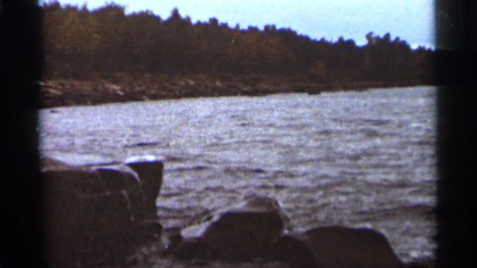 Gentle waves move along a rocky Duluth shoreline with a dark treeline across the water in 1965 8mm footage.