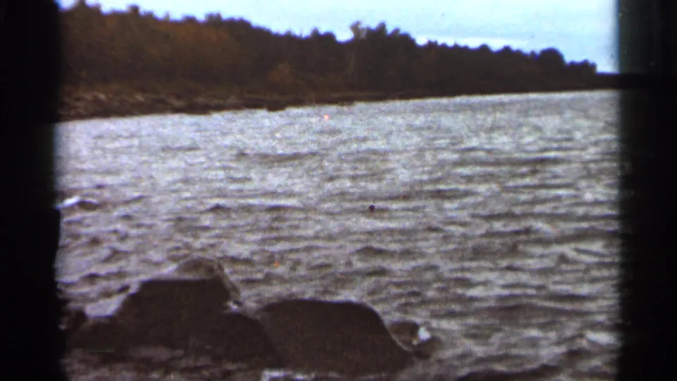 1965 Duluth-area 8mm frame of textured water, low rocks, and a dark tree line across the shore.