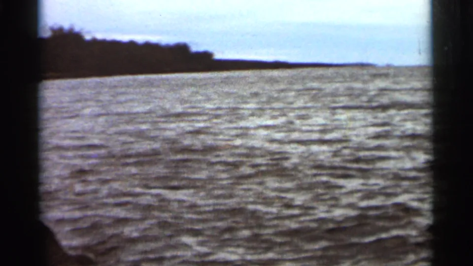 1965 Duluth-area 8mm frame showing open water, a low horizon, and a wooded shoreline tapering at left.