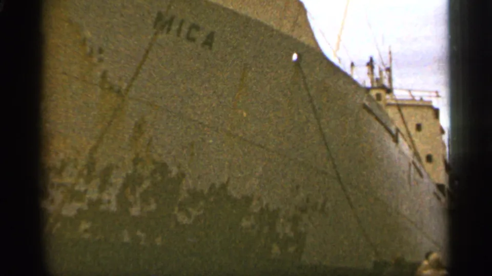1965 Duluth home-movie frame showing the MICA cargo ship's weathered hull, rigging, and distant people near the pier.