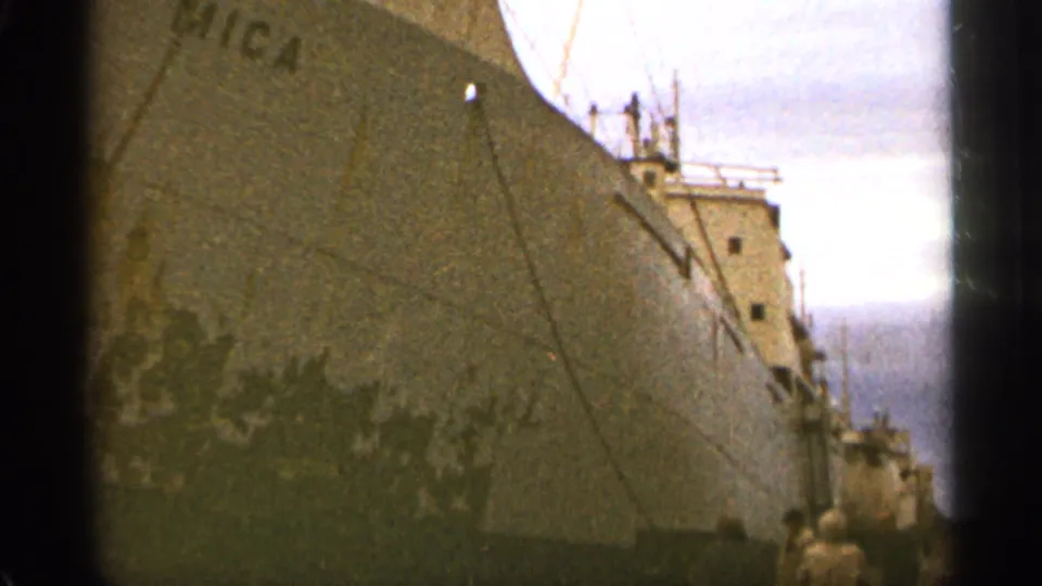 Duluth harbor frame from 1965 showing the MICA cargo ship at dock, with bow, railings, rigging, and distant dockside people.