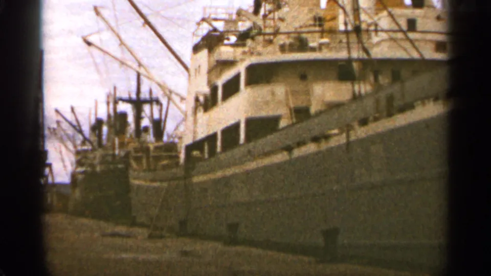 1965 Duluth dockside view with cargo ships, cranes, rigging, and a pier captured from a handheld 8mm perspective.