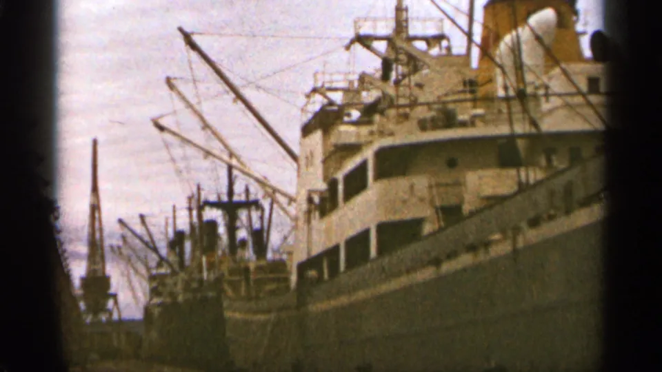 Duluth, Minnesota harbor frame from 1965 showing a docked cargo ship, nearby vessel, cranes, and rigging under daylight.