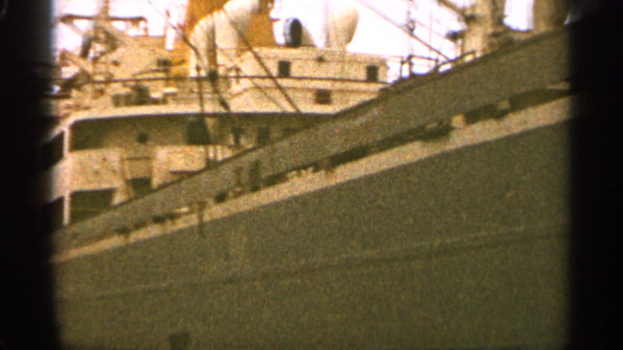 1965 Duluth home-movie frame showing a large docked ship with dark hull, white upper structure, railings, and lifeboats.