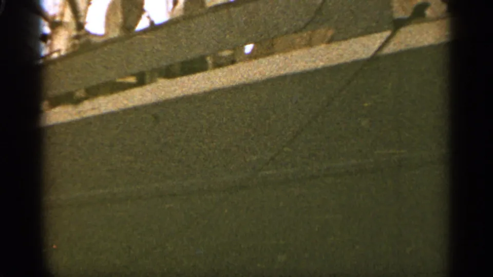 1965 Duluth home-movie frame with the ship's side passing across the view, showing a dark hull, pale railing, and grainy film texture.