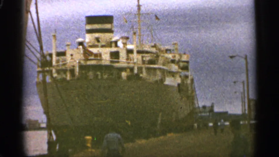 1965 Duluth home-movie frame showing a large docked ship from the pier with cranes, dock lamps, and cloudy sky.