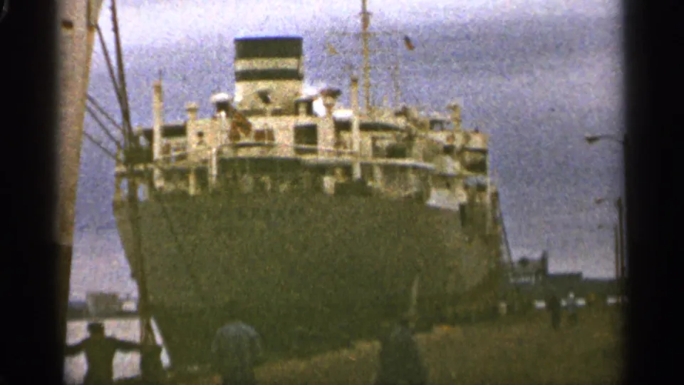 1965 Duluth home-movie frame showing the docked ship's broad hull, upper decks, cranes, and pier-side lamps.