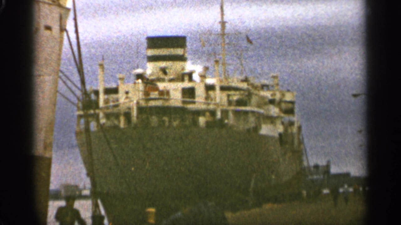Large cargo ship docked at a harbor, viewed from near the stern under an overcast sky.