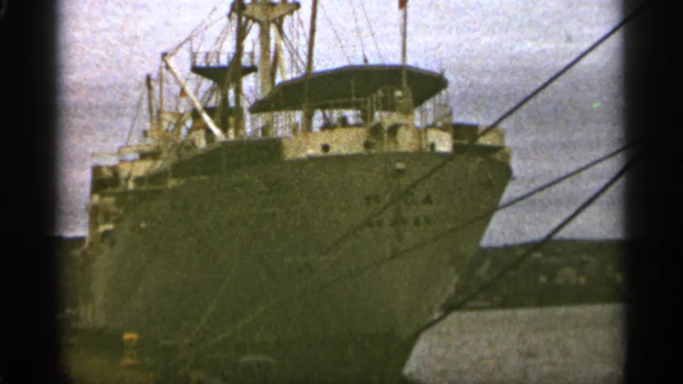 1965 Duluth harbor frame showing the bow and upper deck of a large cargo ship tied up beside the dock.