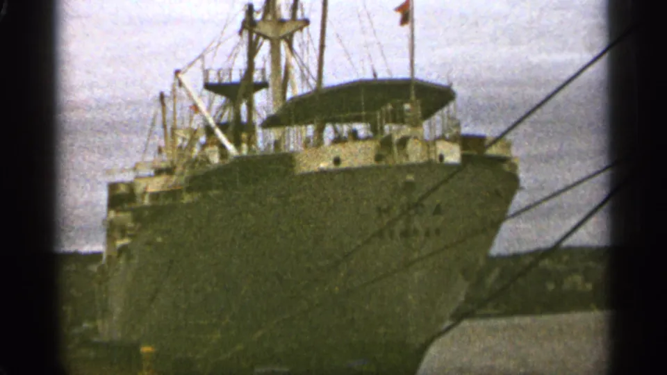 1965 Duluth harbor still of a large cargo ship at dock, with bow, mast rigging, mooring lines, and overcast waterfront background.