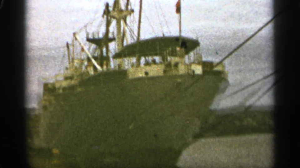 1965 Duluth home-movie frame showing a large cargo ship near a waterway, partly obscured by dark film-frame edges.