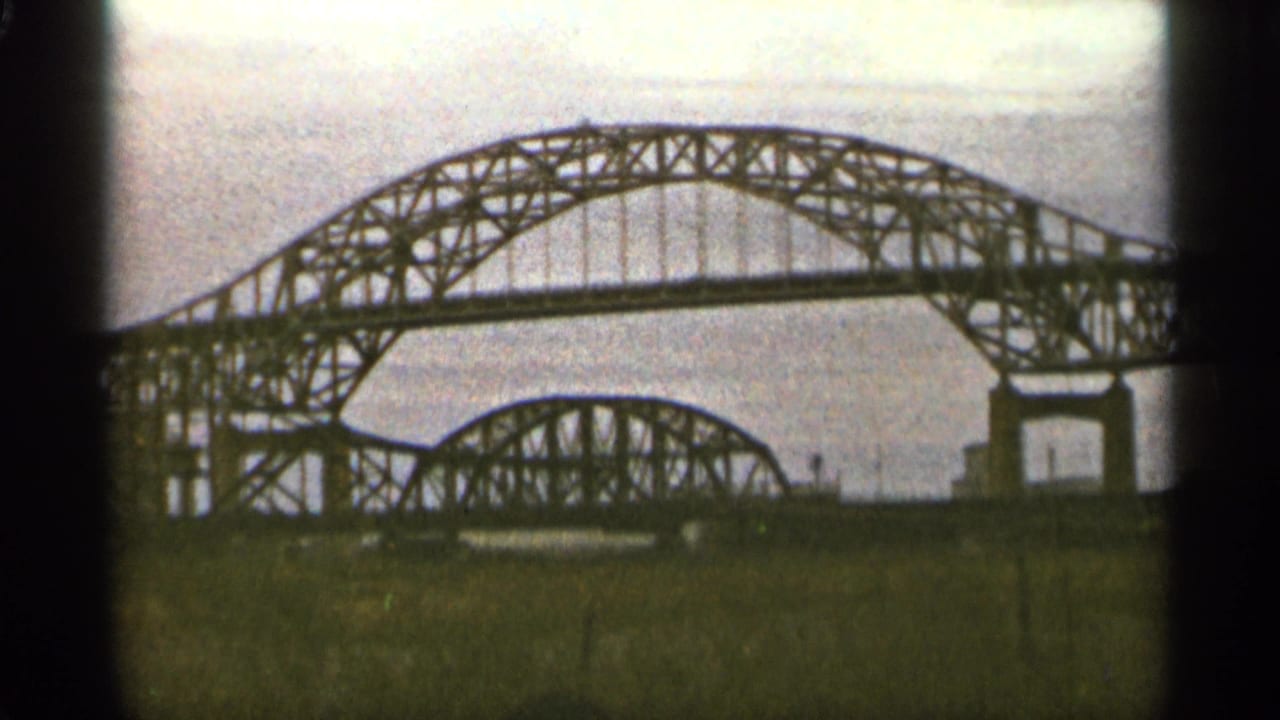 Steel arch bridge seen across water or open ground under a pale sky.