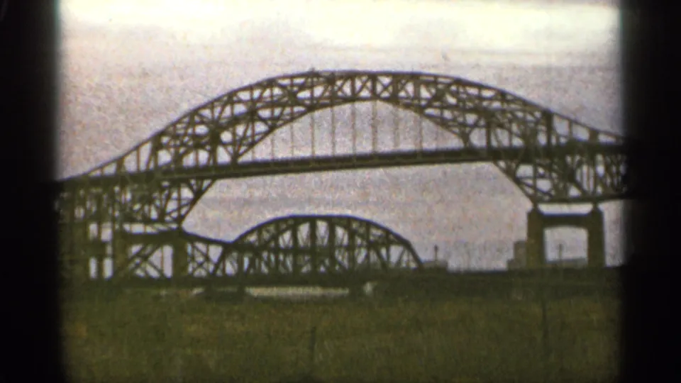 1965 Duluth home-movie view of the Mel Bridge area, with the steel arch span crossing the frame and grass in the foreground.