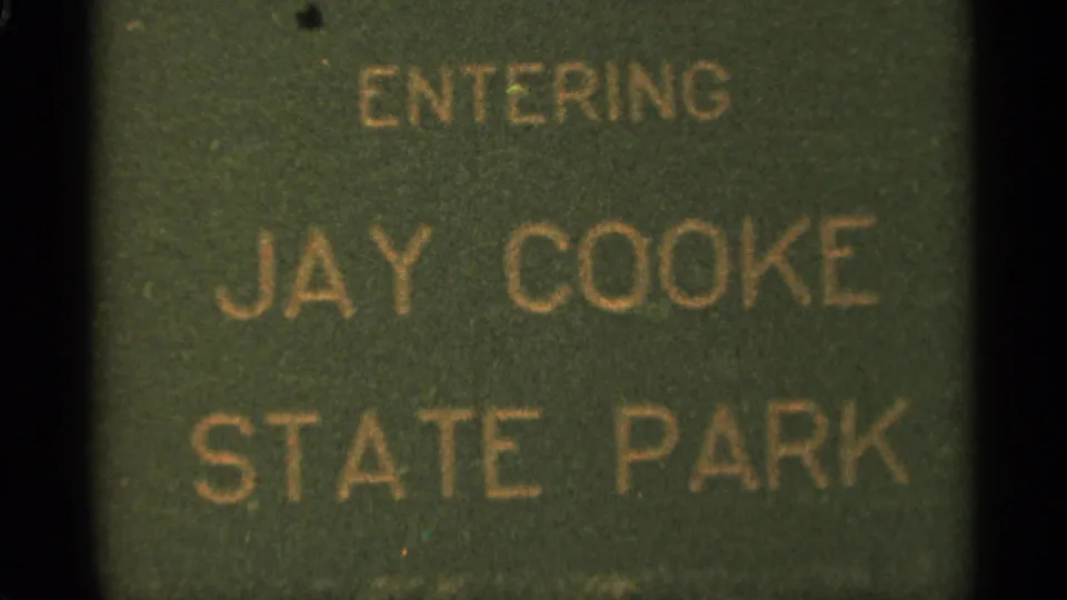 1965 8mm frame from near Duluth showing a green Jay Cooke State Park entry sign with gold lettering and dark film edges.