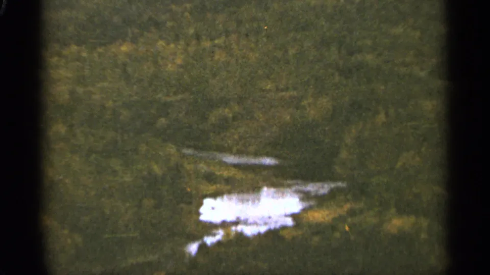 1965 8mm Duluth-area view of a forested landscape and small bright water reflection, framed by dark film borders.