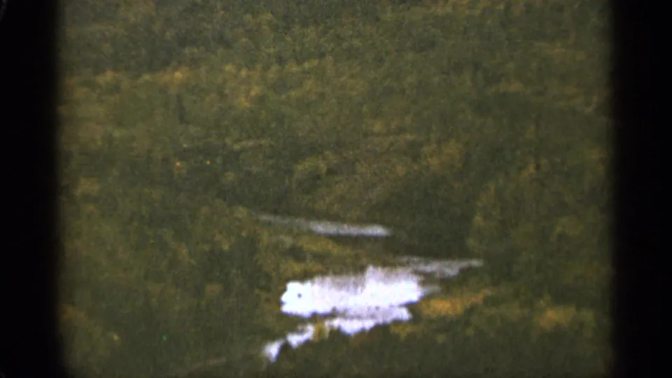 1965 Duluth-area 8mm frame showing a distant wooded slope and reflective water patch with muted color and heavy grain.