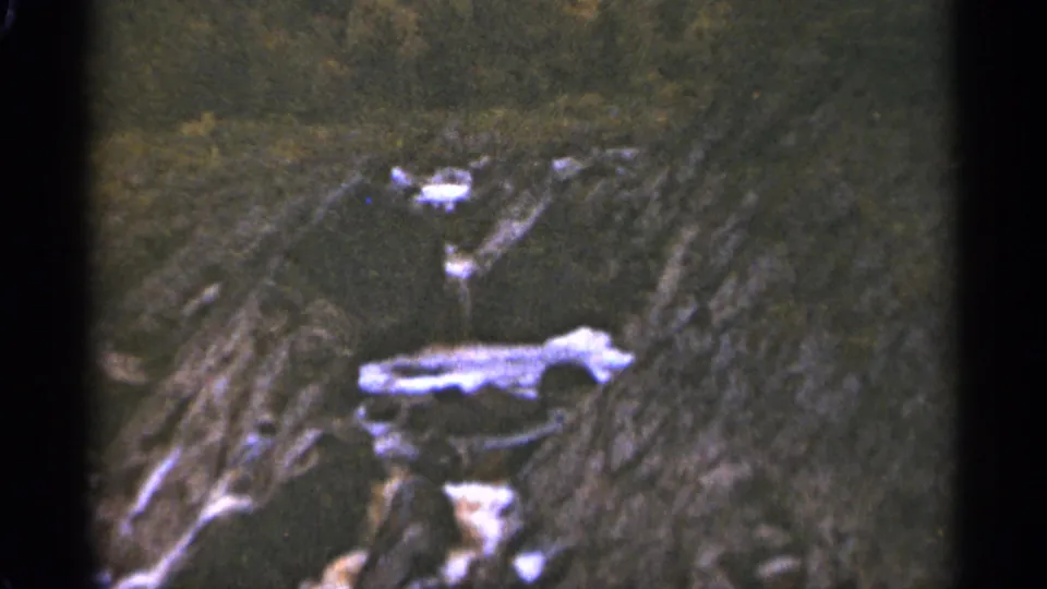 1965 Duluth home-movie frame showing rugged rocky terrain and dark vegetation, with visible 8mm frame edges.