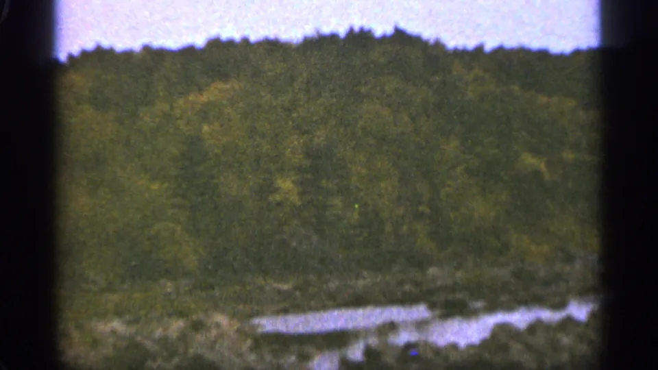 1965 Duluth home-movie frame showing a wooded hillside, uneven shoreline, and small reflective pond in soft 8mm color.