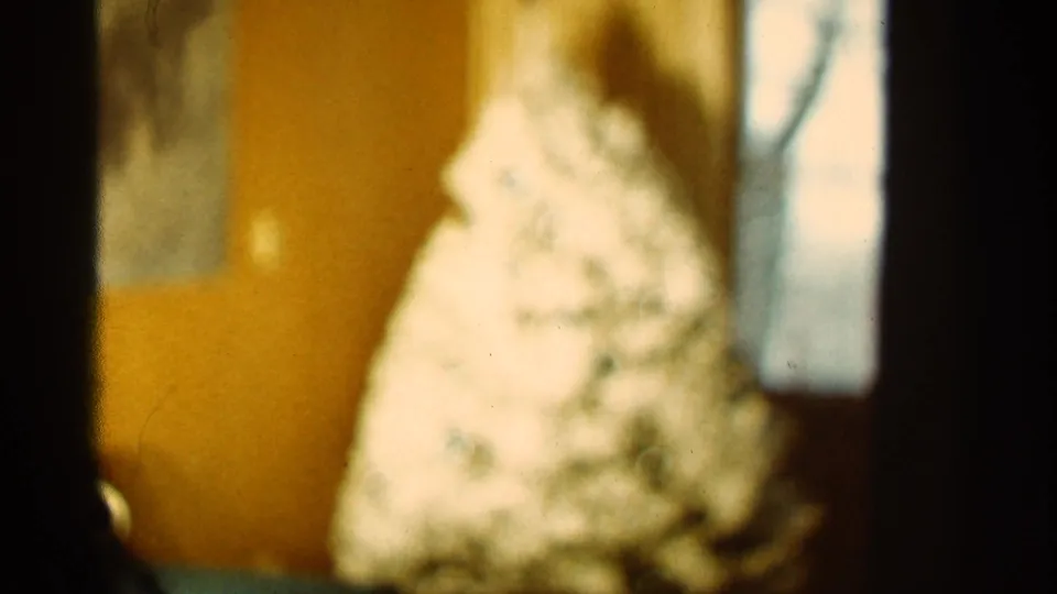 Warm 1965 home-movie frame of a blurred indoor Christmas tree beside a window, framed by dark 8mm edges.