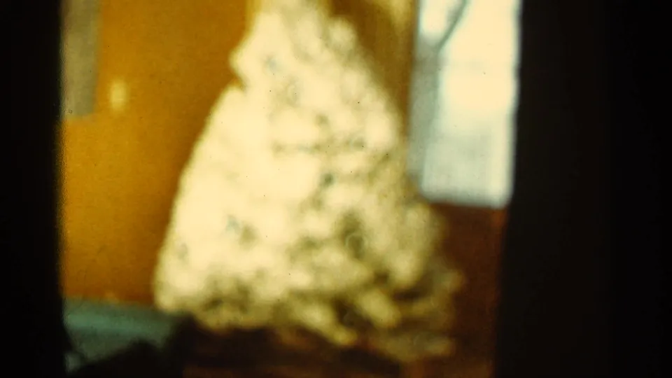 A blurry 1960s 8mm interior view shows a pale cone-shaped object filling the lower room corner, with heavy dark framing at the side.