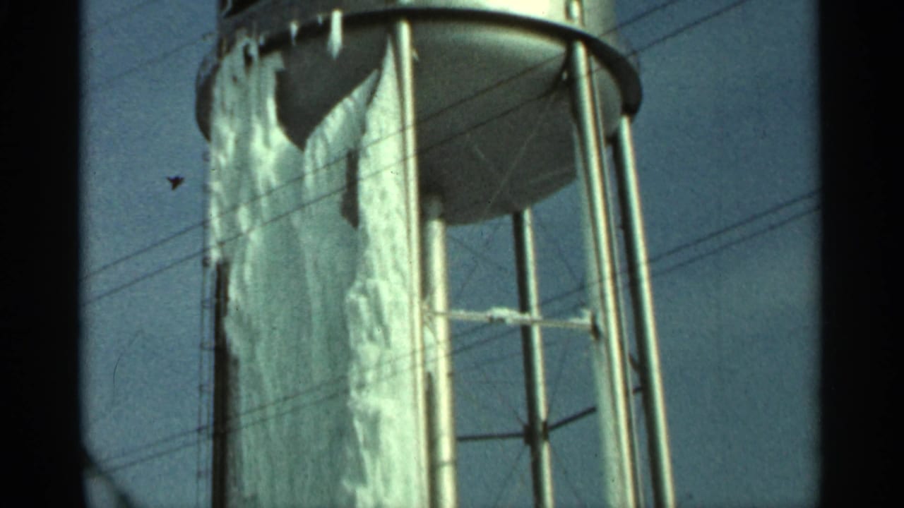 1965 Duluth home-movie frame showing an ice-coated water tank body, steel supports, and utility lines against a pale blue sky.