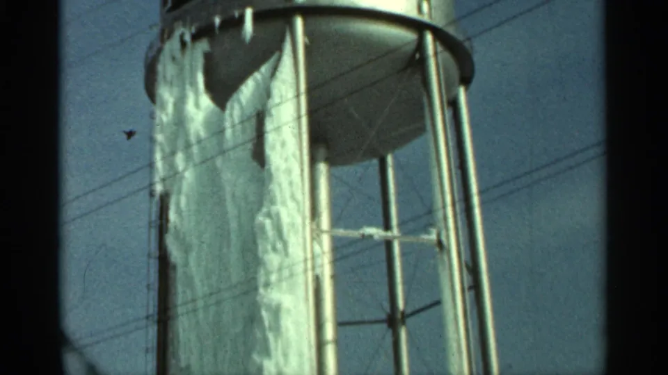 1965 Duluth home-movie frame showing an ice-coated water tank body, steel supports, and utility lines against a pale blue sky.