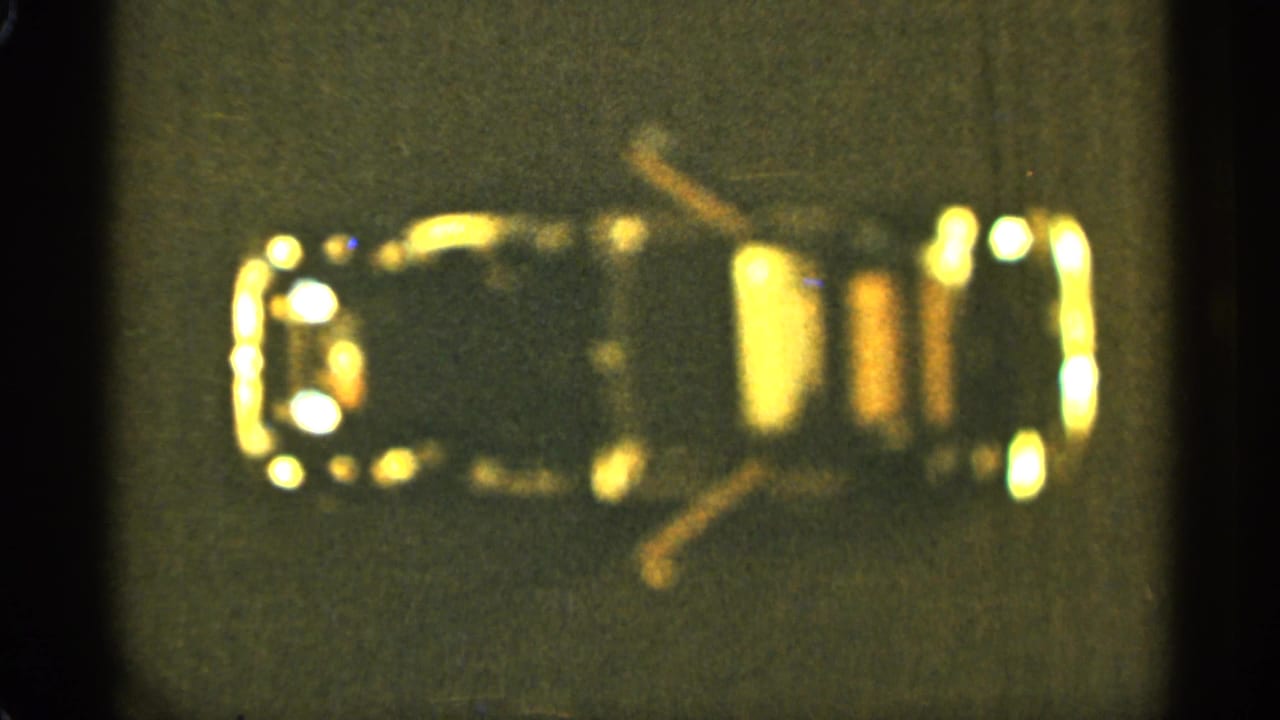 Soft top-down close-up of a small dark object with yellow-gold highlights on a gray surface.