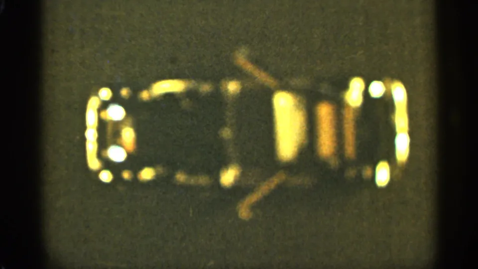 Close-up 8mm frame from Duluth, 1965, with a small glossy object and bright yellow-gold reflections on a gray surface.