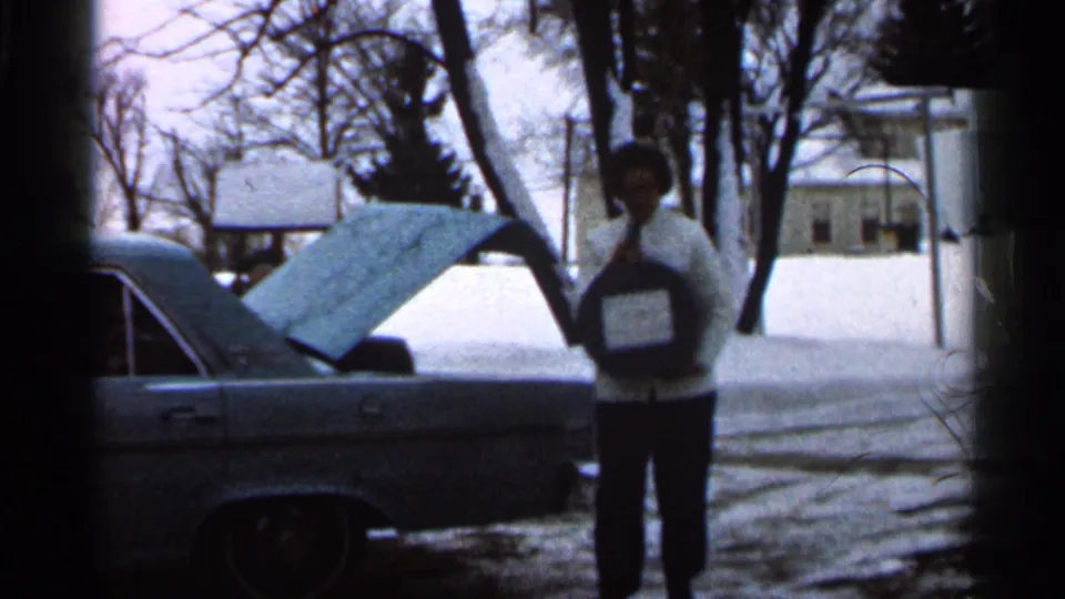 The woman moves from the open trunk holding a round dark object with a white label in a snowy 1966 Eau Claire setting.