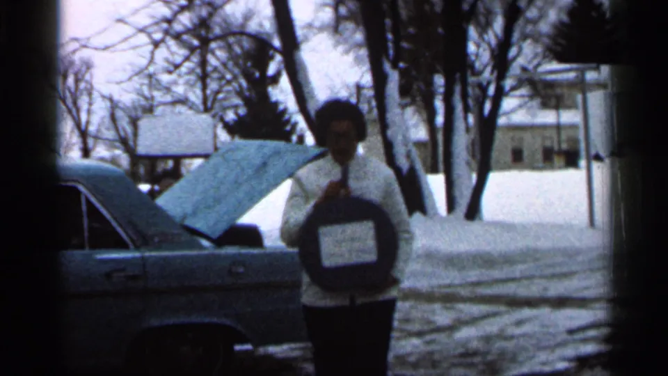 A woman in a light coat approaches the camera with a round labeled object, with the open sedan trunk behind her in snowy Eau Claire.
