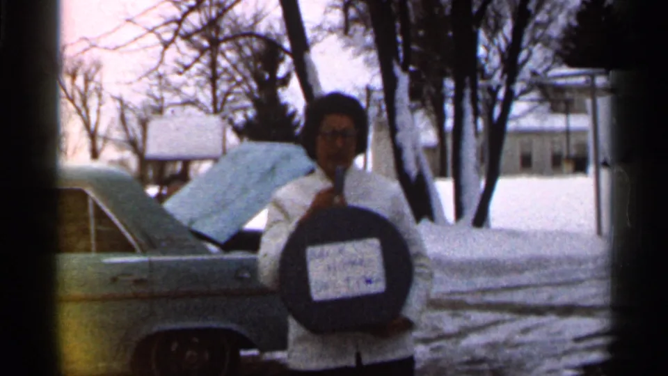 The woman faces the camera holding the round dark labeled object, with the snowy residential street and open car trunk behind her.