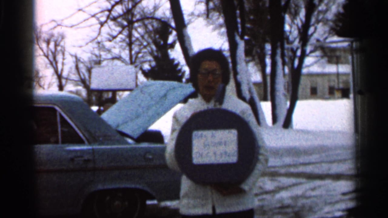 Woman in a white coat stands in snow holding a round sign beside a car with its hood open.