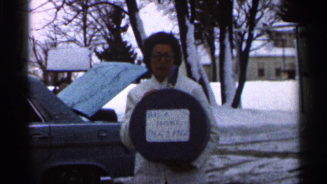 Woman faces the camera with a round sign while standing near a car in a snowy yard.
