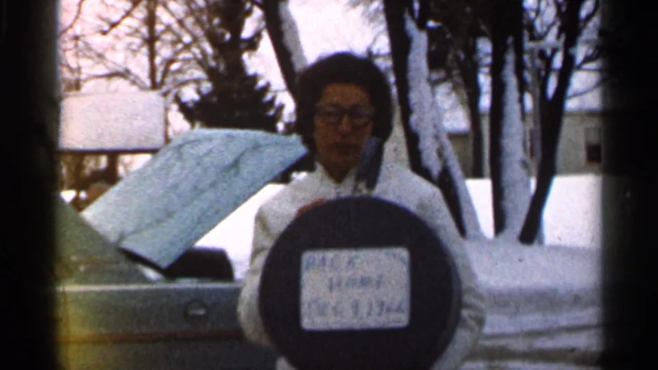 A closer 8mm frame shows the woman and sign in front of snowbanks, with the car hood open and trees behind her.