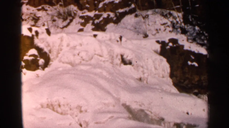 1960s 8mm winter frame showing layered snow drifts over uneven rock, with soft focus and faded home-movie color.