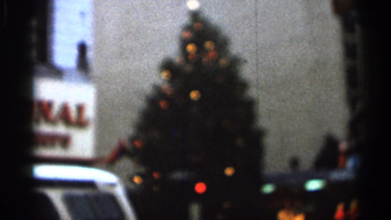 Blurred Christmas tree with colored lights near a partial sign and a white vehicle.