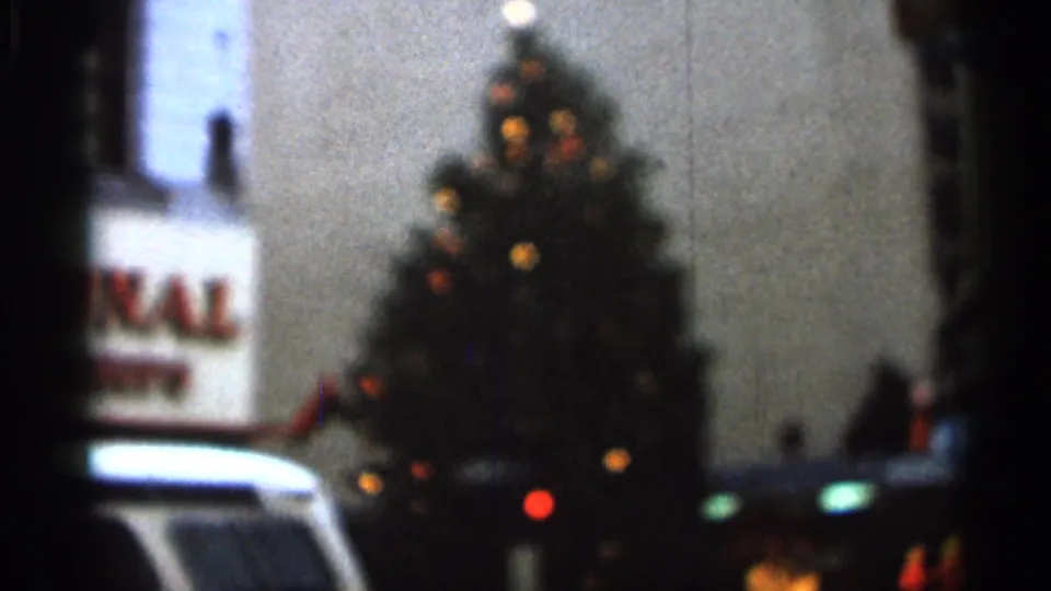 Soft 1966 home-movie view of a lit Christmas tree in a street setting, with a partial sign and white vehicle in the foreground.