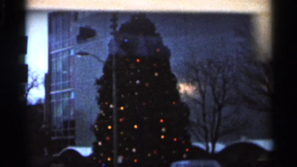 1966 Florida 8mm frame showing a tall illuminated Christmas tree near a modern building and bare trees at dusk or night.