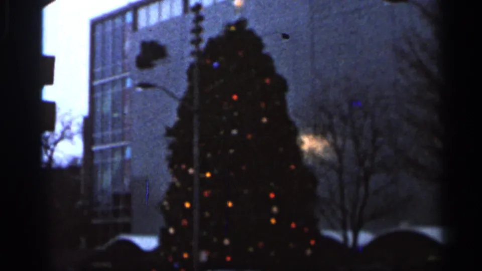 1966 Florida 8mm frame of a lit outdoor Christmas tree against a glass-fronted urban building and winter bare trees.