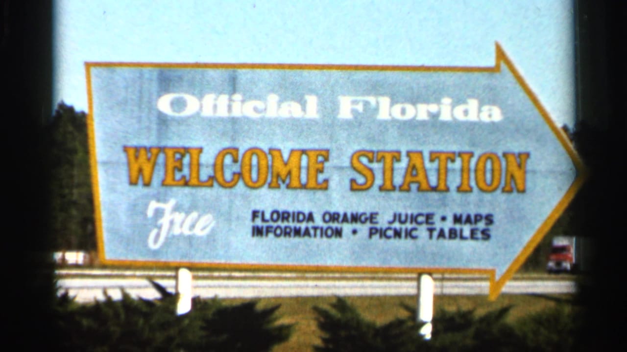 1966 Florida home-movie frame showing an arrow-shaped Official Florida Welcome Station sign advertising free orange juice, maps, and picnic tables.