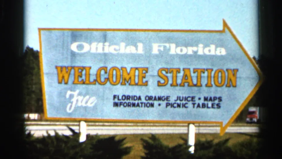 1966 Florida home-movie frame showing an arrow-shaped Official Florida Welcome Station sign advertising free orange juice, maps, and picnic tables.