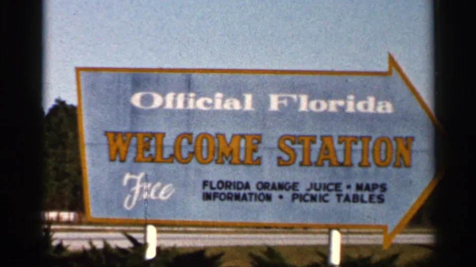 1966 Florida 8mm frame dominated by the state welcome station sign with the arrow point, roadside posts, and dark tree line behind it.