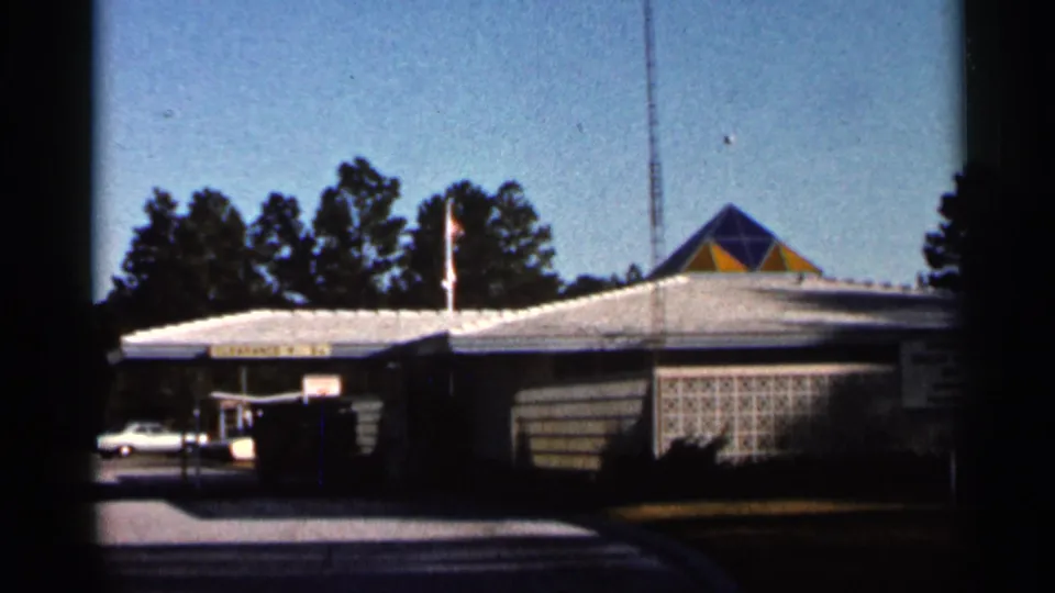 1966 Florida home-movie frame showing a roadside welcome station building with a geometric roof detail, flagpole, parked car, pavement, and trees.