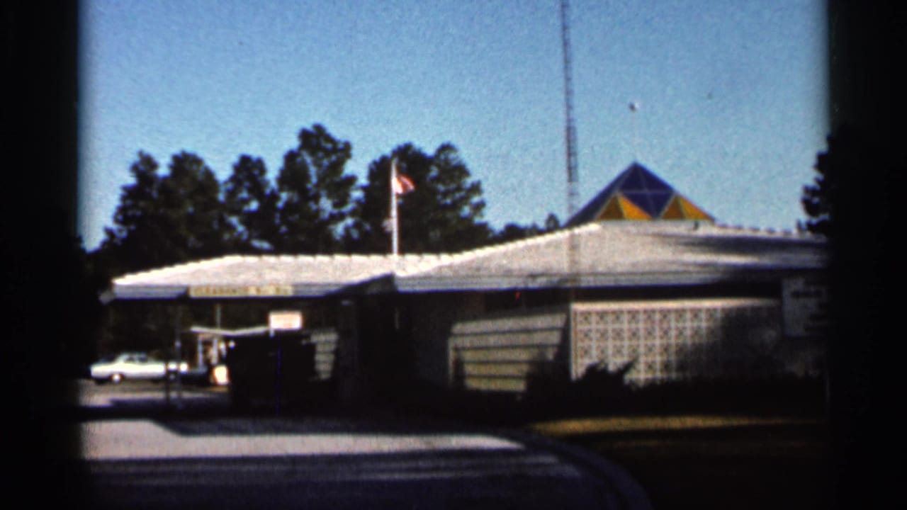 Low commercial building with white roof, flagpole, trees, and a parked white car.