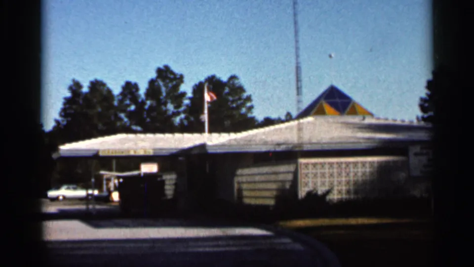 1966 Florida 8mm frame showing a low commercial building with a white roof, patterned wall, flagpole, trees, and parked white car.