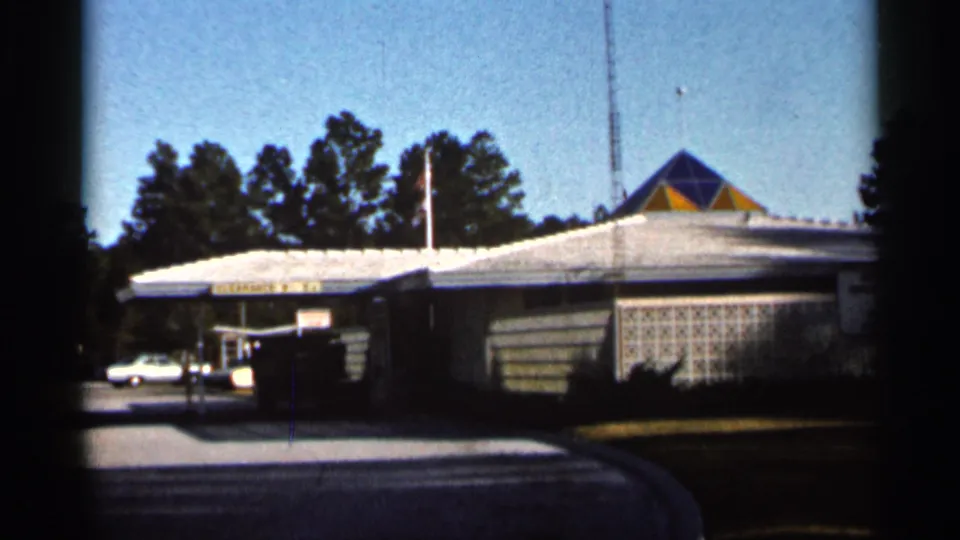 1966 Florida exterior view of a low commercial building with a white roof, geometric roof detail, flagpole, driveway, and trees.